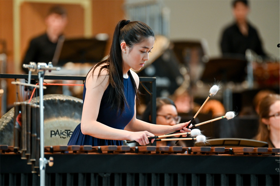 A woman, wearing formal attire, performing on marimba, in an orchestra performance.